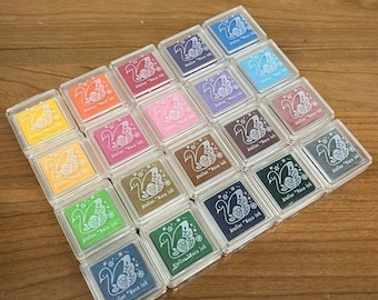 Puede incluir: Una colección de 20 almohadillas de sello de plástico transparente con un diseño de cisne blanco y el texto "Atelier "Naco ink" en cada almohadilla. Las almohadillas están dispuestas en una cuadrícula y se muestran sobre una superficie de madera.