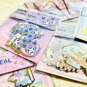 Puede incluir: Un conjunto de paquetes de pegatinas coloridas con varios personajes de la marca Sanrio. Las pegatinas están empaquetadas en bolsas de plástico transparentes con fondos rosa y azul. El texto en las bolsas dice "Kuromi Flake Seal" y "Amorole Flake Seal".