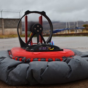 May include: A red and black hovercraft with a large fan and a black skirt. The hovercraft is sitting on a wet concrete surface.