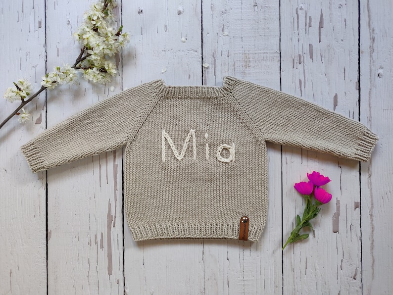 A tan colored first day of the school sweater is laid out on the floor with its arms open. Mia is embroidered on the customized sweater with ecru colored thread. The name engraving on it is embroidered in Latin letters, not in cursive letters.