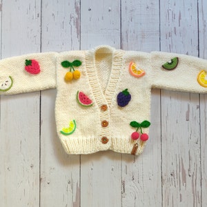 Cream hand-knit chunky cardigan for kids, standing on the floor with arms open. The cardigan features hand-knitted 3D fruits, including a green apple, raspberry, yellow cherry, watermelon, lime, orange, blackberry, red cherry, kiwi, and lemon