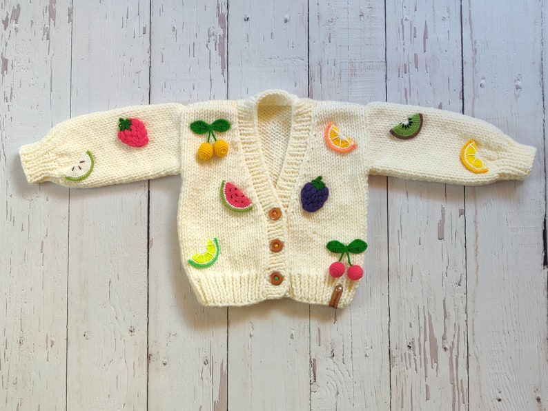 Cream hand-knit chunky cardigan for kids, standing on the floor with arms open. The cardigan features hand-knitted 3D fruits, including a green apple, raspberry, yellow cherry, watermelon, lime, orange, blackberry, red cherry, kiwi, and lemon