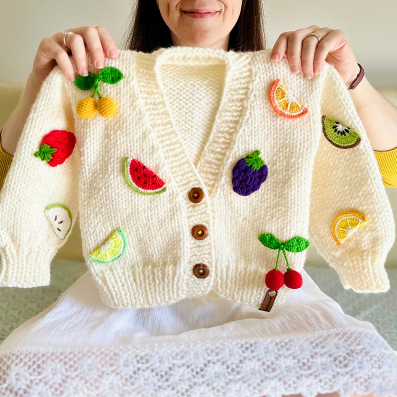 A woman is sitting on a bed, holding a personalized handmade cream-colored children's fruity jacket over her shoulders. This wool cardigan features 10 different crochet fruits on it and can serve as a birthday gift for toddlers who love fruit.