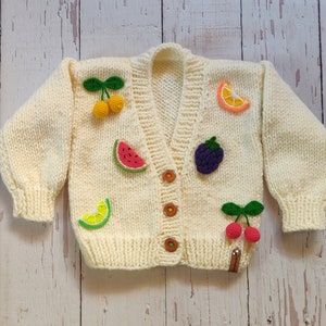 A warm chunky sweater for perfect toddler gift adorned with crochet 3D fruits lies on the floor.