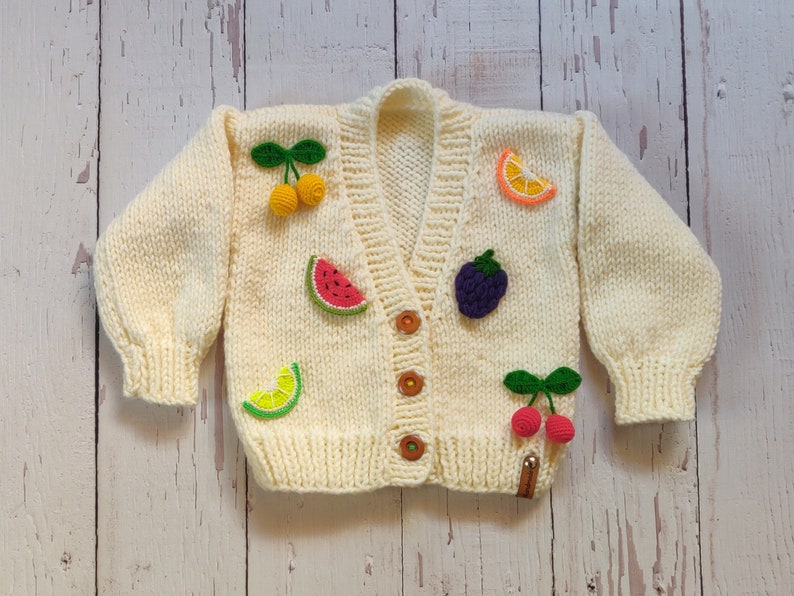 A warm chunky sweater for perfect toddler gift adorned with crochet 3D fruits lies on the floor.