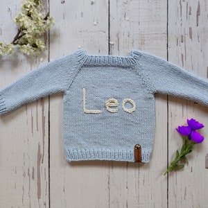 A light blue colored hand knit birthday jumper for toddler is laid out on the floor with its arms open. Leo is embroidered on the handmade kids jumper with ecru colored thread.