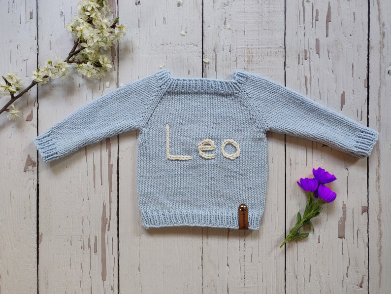 A light blue colored hand knit birthday jumper for toddler is laid out on the floor with its arms open. Leo is embroidered on the handmade kids jumper with ecru colored thread.