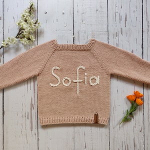 A pastel peach colored personalized handmade sweater with name embroidered for baby is laid out on the floor with its arms open. Sofia is embroidered on the sweater with ecru colored cotton thread.