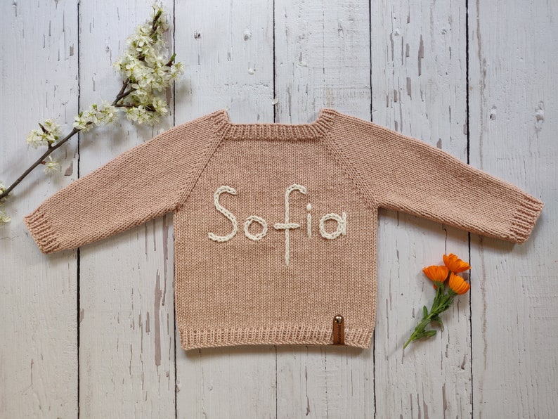 A pastel peach colored personalized handmade sweater with name embroidered for baby is laid out on the floor with its arms open. Sofia is embroidered on the sweater with ecru colored cotton thread.