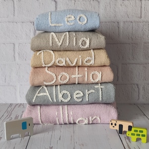 Monogram name embroidered kids sweaters from newborn to 6 years old are folded and stacked on the floor. The names on the sweaters in the photo are written in ecru. All sweaters are in different colors, but all have names embroidered in ecru thread.