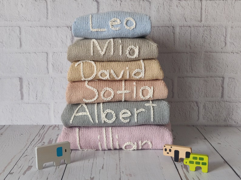 Monogram name embroidered kids sweaters from newborn to 6 years old are folded and stacked on the floor. The names on the sweaters in the photo are written in ecru. All sweaters are in different colors, but all have names embroidered in ecru thread.