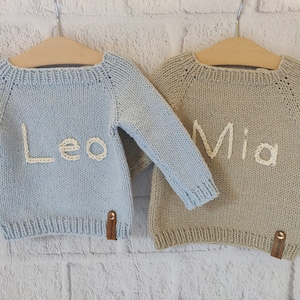 2 newborn keepsake gift sweaters are hanging on the clothes hanger. Leo is embroidered with ecru color thread on the light blue personalized sweater on the left. Mia is embroidered with ecru color on the tan color custom knit sweater on the right.