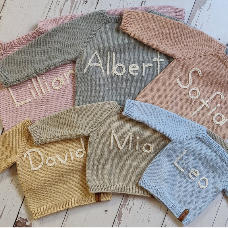 Hand embroidered baby name sweaters are lying on the floor. The name Lilian is embroidered on the hand knitted lavender sweater, Albert on the gray, Sofia on the pastel peach, David on the beige, Mia on the tan, and Leo on the light blue.