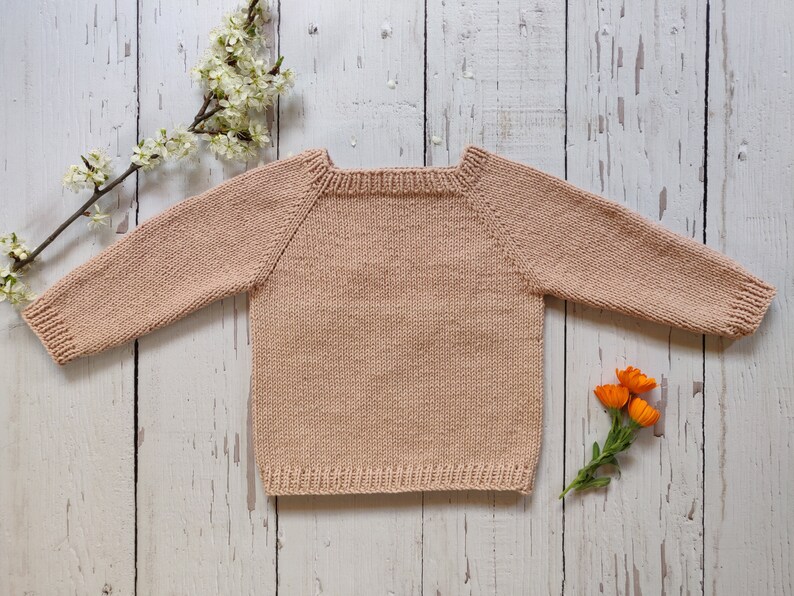 Back of a pastel peach colored hand-knitted sweater is laid out on the floor with its arms open.