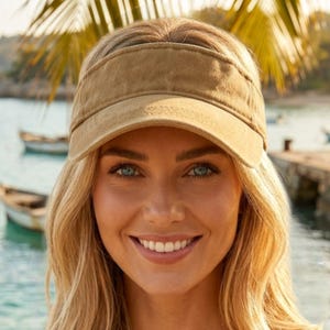 May include: A tan visor with a curved peak. The visor is made of a soft, textured fabric. The background shows a tropical scene with boats and water. The visor is worn by a person with blonde hair and blue eyes.
