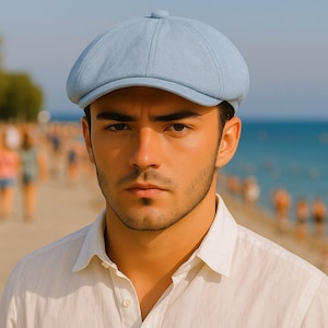 May include: A light blue newsboy cap with a rounded crown and a small, stiff brim. The cap is made of a textured fabric and is worn by a person wearing a white button-down shirt. The background shows a beach and ocean.