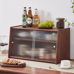 May include: A wooden cabinet with glass doors and shelves. The cabinet is filled with various items, including tea bags, plates, and bottles. The cabinet has a brown finish and gold-colored hardware.