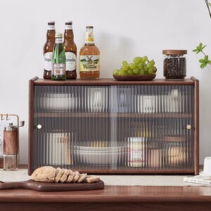 May include: A brown wooden cabinet with two glass doors. The cabinet has two shelves and is filled with various items, including plates, bowls, jars, and bottles. There are also some grapes in a bowl on top of the cabinet.