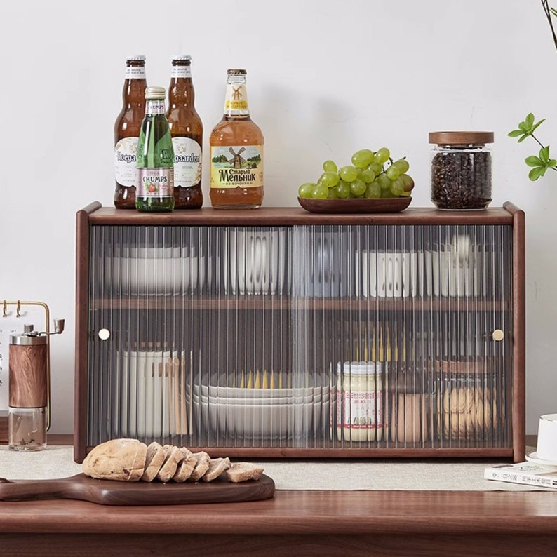May include: A brown wooden cabinet with two glass doors. The cabinet has two shelves and is filled with various items, including plates, bowls, jars, and bottles. There are also some grapes in a bowl on top of the cabinet.