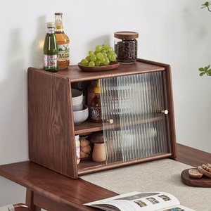 May include: A dark brown wooden cabinet with a glass door and two shelves. The cabinet has a gold knob and is filled with various items, including bottles, jars, and bowls. There are grapes on a plate on top of the cabinet.