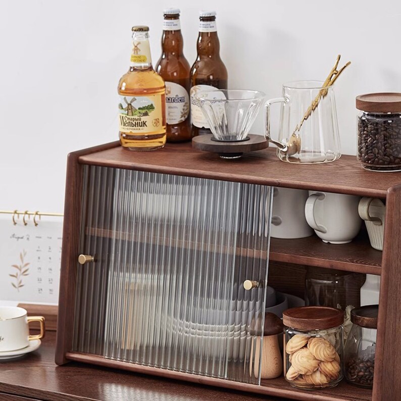 May include: A wooden cabinet with a glass door and two shelves. The cabinet is filled with various items, including bottles of beer, a coffee filter, a glass pitcher, and a jar of coffee beans. The cabinet has a brown wood finish and gold-colored handles.