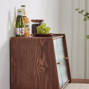 May include: A wooden cabinet with a glass door and two brass knobs. The cabinet is sitting on a wooden table and has a small plate of grapes on top. There are two bottles of beverage on the top shelf.