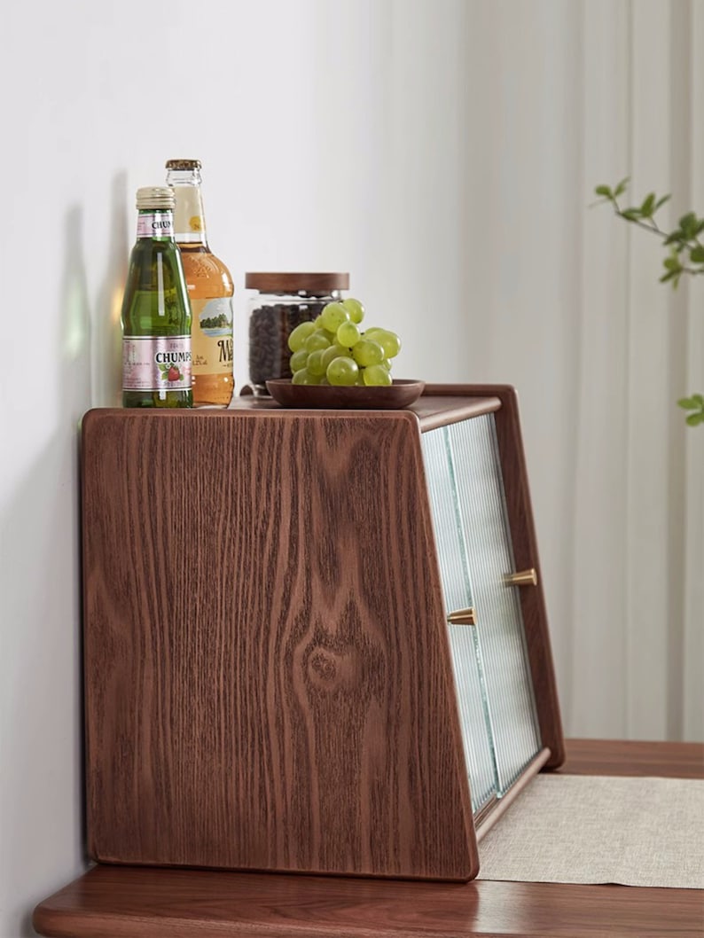 May include: A wooden cabinet with a glass door and two brass knobs. The cabinet is sitting on a wooden table and has a small plate of grapes on top. There are two bottles of beverage on the top shelf.