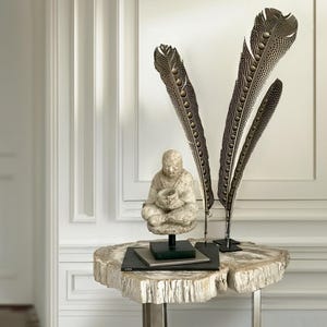 Può includere: Una scultura in pietra bianca di una figura in meditazione si trova su un tavolo in legno pietrificato bianco con due grandi piume marroni e nere in supporti decorativi.