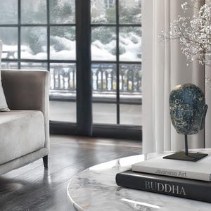 Peut inclure: Une sculpture de tête de Bouddha en pierre grise sur un support noir, avec deux livres sur une table en marbre blanc. Le livre du haut porte le texte "Javanese Art" et le livre du bas porte le texte "BUDDHA".