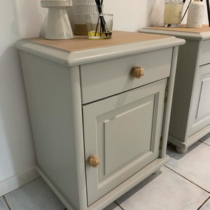 May include: Two matching bedside tables with a light grey painted finish. Each table has a drawer with a rope knob and a cabinet door with a rope knob. The top of the table is a light wood colour.