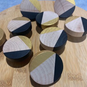 May include: Nine round wooden knobs with a geometric design. Each knob is painted in black, gold, and natural wood tones. The knobs are arranged in a circular pattern on a wooden surface.