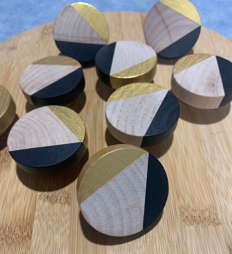 May include: Nine round wooden knobs with a geometric design. Each knob is painted in black, gold, and natural wood tones. The knobs are arranged in a circular pattern on a wooden surface.