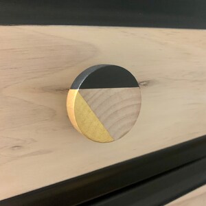 May include: A round wooden drawer knob with a black and gold geometric design. The knob is made of natural wood with a black painted section and a gold painted section.