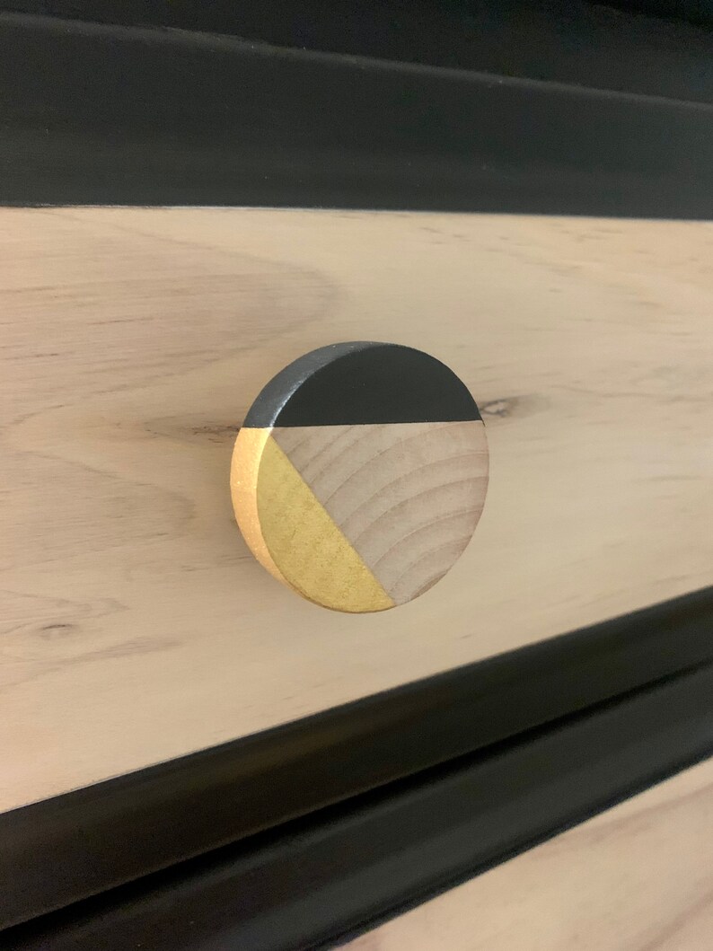 May include: A round wooden drawer knob with a black and gold geometric design. The knob is made of natural wood with a black painted section and a gold painted section.