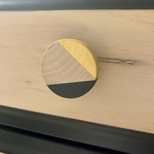 May include: A round wooden drawer knob with a black and gold geometric design. The knob is made of natural wood with a black painted bottom and a gold painted top.