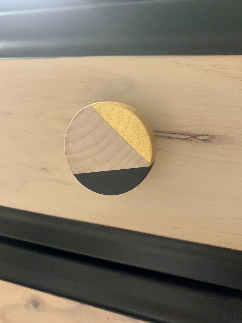 May include: A round wooden drawer knob with a black and gold geometric design. The knob is made of natural wood with a black painted bottom and a gold painted top.