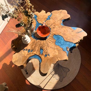 Op de afbeelding: Een unieke salontafel gemaakt van een grote houten plaat met blauwe hars die de scheuren vult. De tafel heeft een natuurlijke houtkleur en wordt ondersteund door drie zwarte metalen haarspeldpoten. De tafel staat op een rond jutekleed met een decoratief ontwerp.
