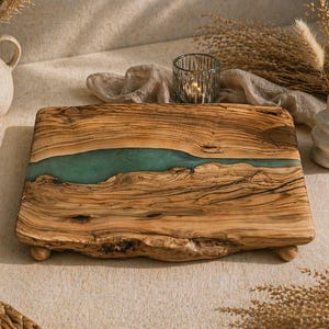 May include: A wooden serving board with a teal resin river design. The board has a natural wood grain pattern and is supported by small wooden feet. Decorative vases and a candle holder are in the background.