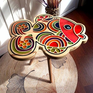 May include: A wooden side table shaped like a colorful fish with a geometric design. The table has three legs and is sitting on a woven rug with a ginkgo leaf design.