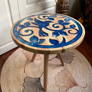 May include: A round wooden side table with a blue and white resin inlay featuring a floral design. The table has three wooden legs and is sitting on a woven rug.