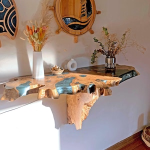 Può includere: Un ripiano a muro in legno con un design a bordo vivo. Il ripiano è realizzato in legno naturale con un inserto in resina epossidica blu. Il ripiano è montato a parete e ha un aspetto rustico e naturale.