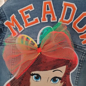 Puede incluir: Una chaqueta vaquera azul con un gran parche de fieltro naranja que dice "MEADOW" y un parche de lentejuelas rojas, verdes y amarillas de Ariel de La Sirenita con un gran lazo rojo.