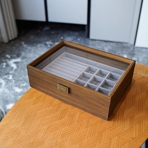 Puede incluir: Una caja de joyería rectangular de madera con una tapa de vidrio transparente. El interior presenta compartimentos de terciopelo gris para anillos y otras joyas. La caja se encuentra sobre una mesa naranja estampada, mostrando un diseño clásico.