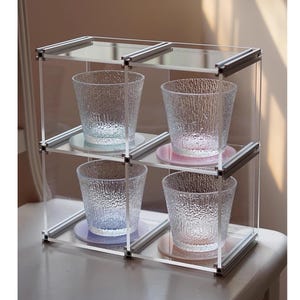 Puede incluir: Una vitrina transparente de tres niveles con cuatro vasos de vidrio texturizado. Cada vaso se asienta sobre un posavasos de color: azul, rosa y morado. La vitrina tiene un marco plateado y estantes transparentes.