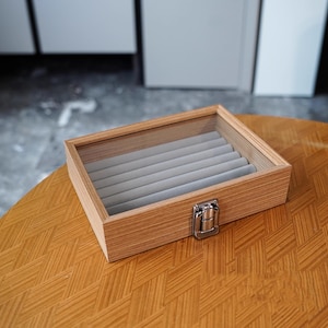 May include: A wooden jewelry box with a clear glass top and a silver-colored clasp. The interior features gray velvet-lined compartments. The box is sitting on a wooden table with a geometric pattern.