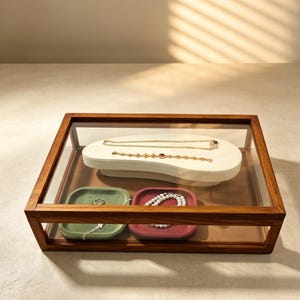 May include: A rectangular wooden display box with a glass lid containing a white kidney-shaped jewellery stand holding two gold chains, plus a green square tray with a silver ring and a red square tray holding a white pearl necklace.
