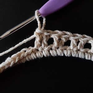 May include: Close-up of a crocheted piece in progress, showing a row of single crochet stitches in beige yarn. A purple crochet hook is visible in the foreground.