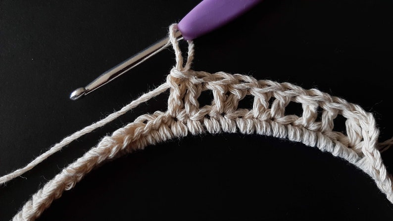 May include: Close-up of a crocheted piece in progress, showing a row of single crochet stitches in beige yarn. A purple crochet hook is visible in the foreground.