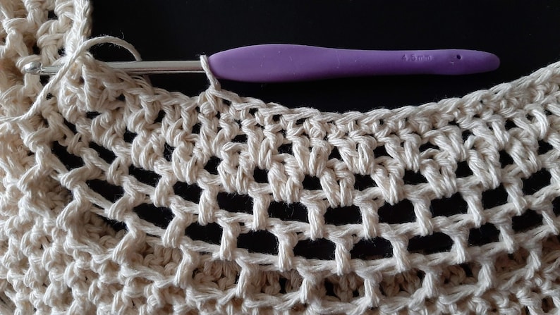 May include: Close-up of a crocheted piece in a cream color with a purple crochet hook resting on top. The crochet hook is 4.5 mm.