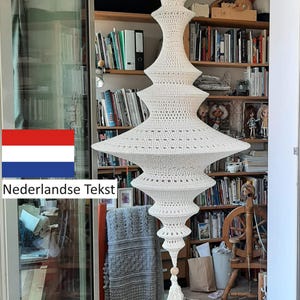 Op de afbeelding: Een witte gecrochete macramé hanglamp met een kwast. De lamp hangt aan het plafond in een kamer met boekenkasten en een spinnewiel.